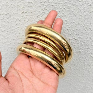 May include: A set of three stacked gold-toned bangles with a smooth, polished, and reflective surface. The bracelets are circular, thick, and feature a rounded, tube-like profile. The metal has a bright, warm yellow finish that catches the light.