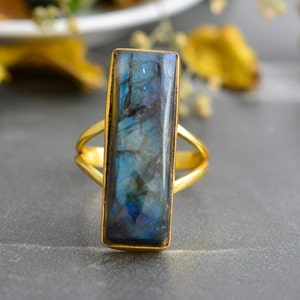 Könnte beinhalten: Ein goldener Ring mit einem großen, rechteckigen, schillernden Labradorit-Edelstein. Der Stein hat eine blaue und grüne Farbe mit goldenen Schimmern.