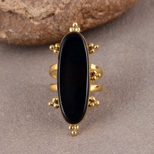 Peut inclure: Une bague en or avec une grande pierre ovale noire. La bague a un design décoratif avec de petites perles dorées autour de la pierre.