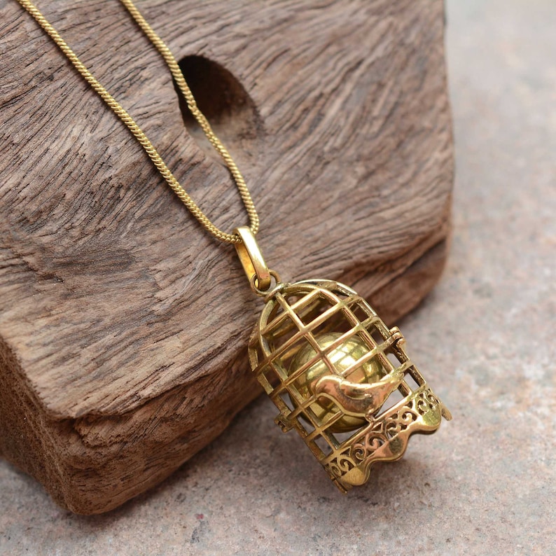 Puede incluir: Un collar de tono dorado con un colgante de jaula que presenta un peque&ntilde;o p&aacute;jaro en su interior. La jaula est&aacute; abierta por un lado y tiene un dise&ntilde;o delicado.