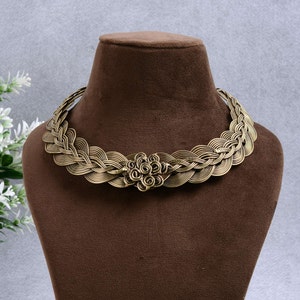 May include: A gold-toned metal necklace with a braided design and a floral centerpiece. The necklace is displayed on a brown velvet mannequin.