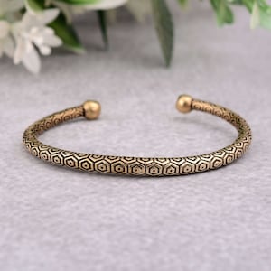 May include: A gold-toned metal bracelet with a geometric pattern. The bracelet is open and has a rounded end.