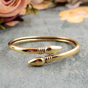 May include: A gold-toned bangle bracelet with two pointed ends. The bracelet is made of a thick, smooth metal and has a simple, elegant design.