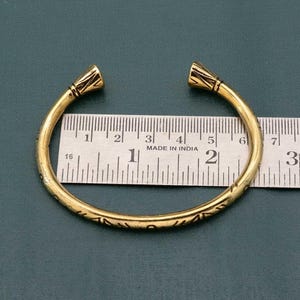 May include: A gold-toned bangle bracelet with a textured design. The bracelet is approximately 2 inches in diameter and is measured against a ruler.