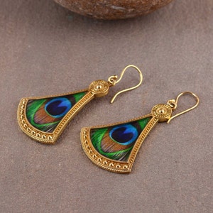 Puede incluir: Un par de pendientes colgantes de tono dorado con un diseño de pluma de pavo real. Los pendientes están hechos de metal y tienen un borde de cuentas.