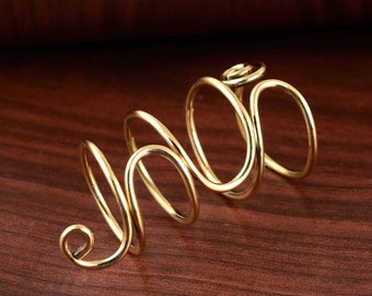 May include: A gold-colored, abstract ring with a swirling, open design. The ring features a continuous, looping form, creating an elegant and modern aesthetic. The jewelry piece is set against a warm, brown wooden surface.