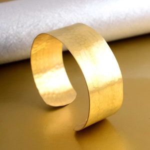 Puede incluir: Un brazalete ancho de metal martillado de color dorado. El brazalete está hecho de una pieza gruesa y plana de metal que ha sido martillada para crear una superficie texturizada.