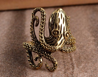 Può includere: Un anello a forma di polpo dorato con un design testurizzato dettagliato. L'anello presenta un tentacolo arrotolato sul lato e una testa di polpo testurizzata con gli occhi.