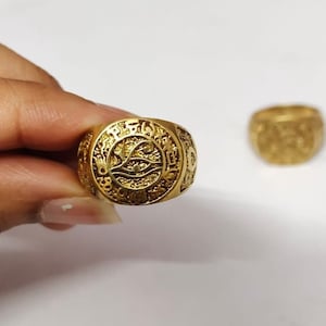 May include: A gold ring with an intricate design featuring an eye and other symbols. The ring is worn on a finger.