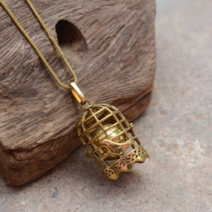 Puede incluir: Un collar de tono dorado con un colgante de jaula de p&aacute;jaros. La jaula est&aacute; abierta y un p&aacute;jaro peque&ntilde;o est&aacute; dentro. La jaula de p&aacute;jaros est&aacute; detallada con dise&ntilde;os intrincados.