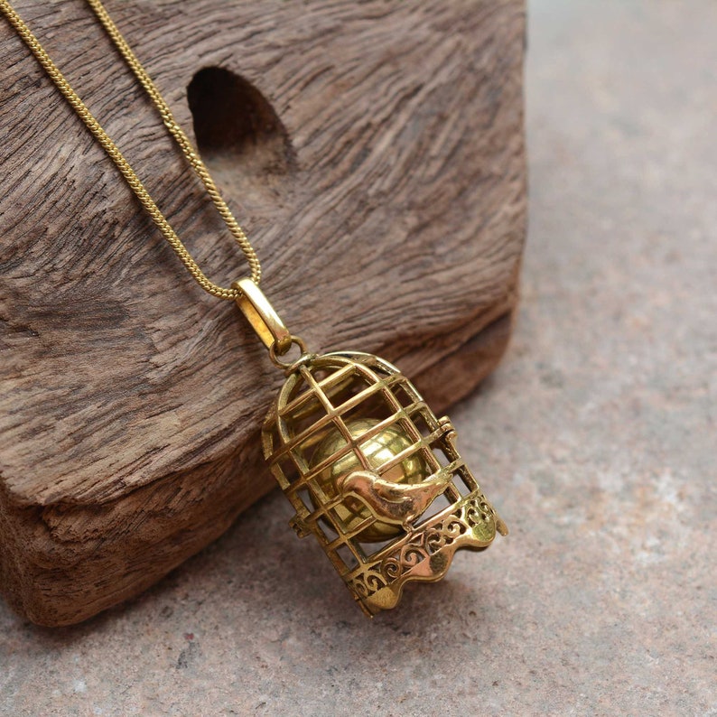 Puede incluir: Un collar de tono dorado con un colgante de jaula de p&aacute;jaros. La jaula est&aacute; abierta y un p&aacute;jaro peque&ntilde;o est&aacute; dentro. La jaula de p&aacute;jaros est&aacute; detallada con dise&ntilde;os intrincados.