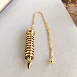 Op de afbeelding: Een gouden metalen slinger met een puntige punt en een ketting. De slinger heeft een cilindrisch lichaam met een reeks horizontale groeven.