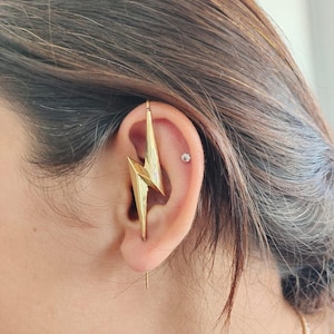 May include: Gold lightning bolt ear cuff with a small clear gemstone stud in the earlobe.