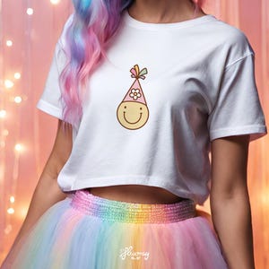 May include: A white cropped t-shirt with a smiling cartoon face wearing a pink party hat with a flower on top. The shirt is being worn by a person with rainbow colored hair and a rainbow tulle skirt.