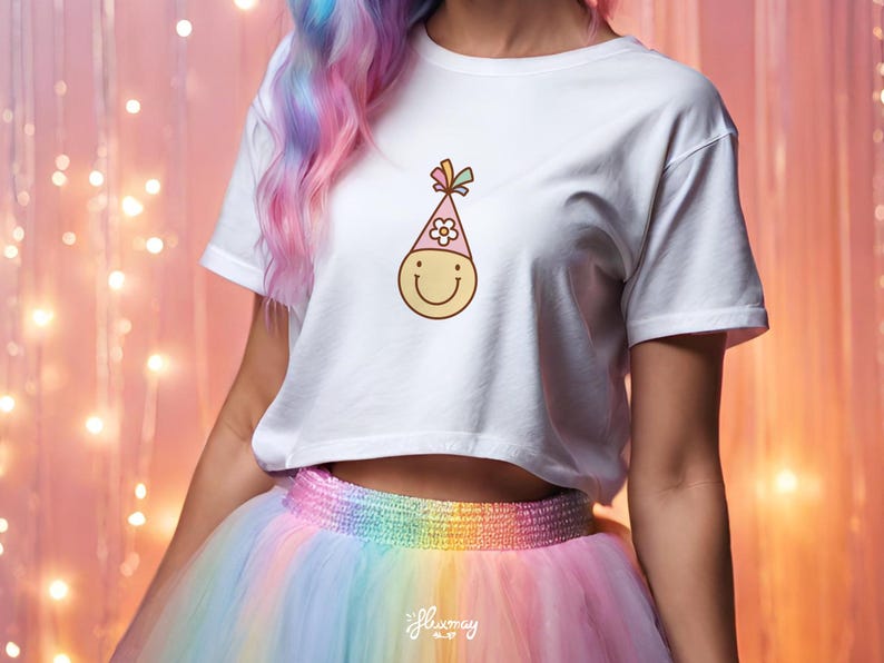 May include: A white cropped t-shirt with a smiling cartoon face wearing a pink party hat with a flower on top. The shirt is being worn by a person with rainbow colored hair and a rainbow tulle skirt.