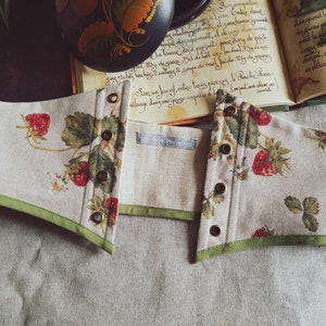 May include: A beige corset with a strawberry pattern, featuring red strawberries and green leaves. The corset has a green trim and bronze eyelets. The fabric is a linen blend. The corset is open in the front.