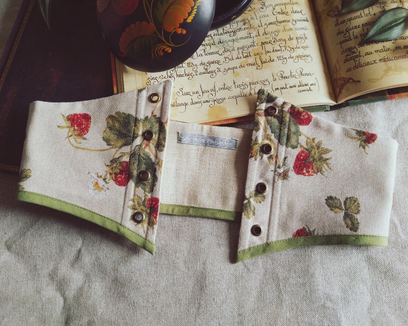 May include: A beige corset with a strawberry pattern, featuring red strawberries and green leaves. The corset has a green trim and bronze eyelets. The fabric is a linen blend. The corset is open in the front.