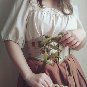 May include: A cream-colored blouse with lace detailing and puffed sleeves is paired with a strawberry-print corset and a brown skirt. The corset features green ribbon lacing. A straw hat is held in the hands.