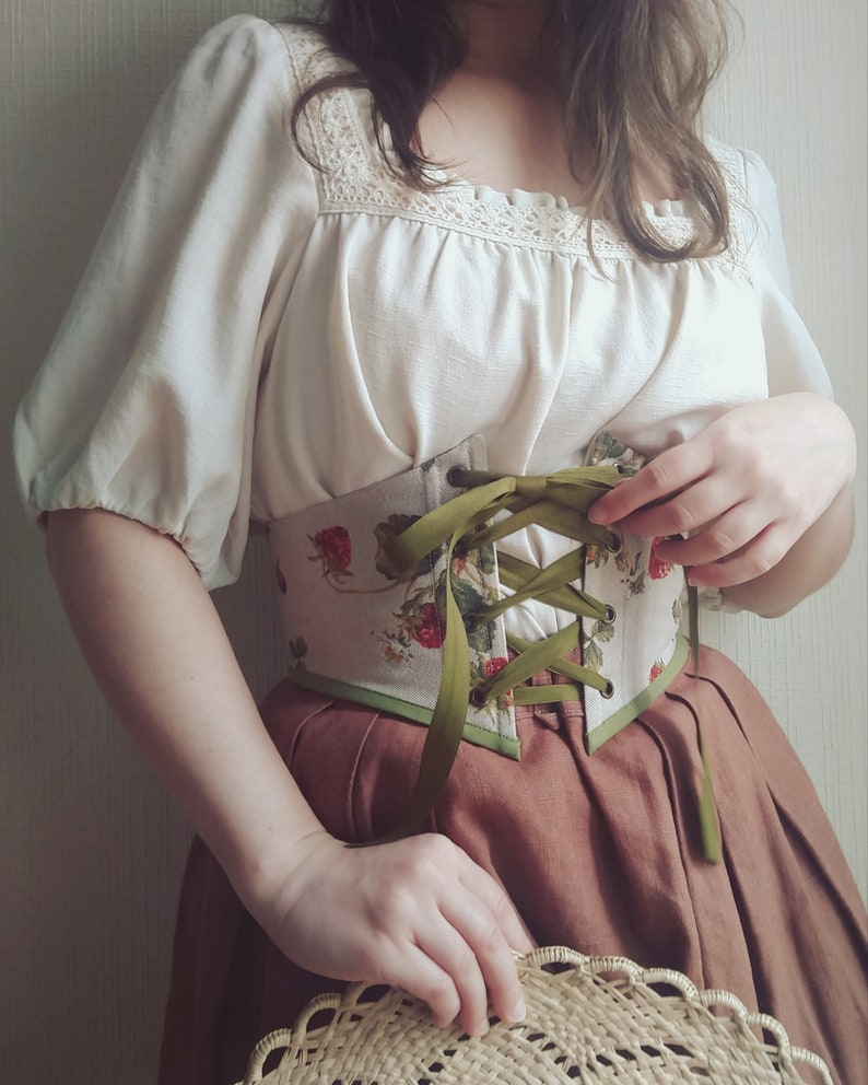 May include: A cream-colored blouse with lace detailing and puffed sleeves is paired with a strawberry-print corset and a brown skirt. The corset features green ribbon lacing. A straw hat is held in the hands.