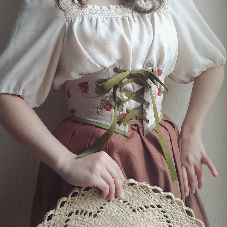 May include: A cream-colored blouse with lace trim and puffed sleeves is paired with a brown skirt. A strawberry-printed corset with green ribbon lacing is the focal point. A straw hat is held in the hands.