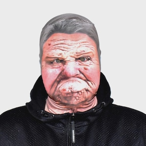 Könnte beinhalten: Eine Vollmaske mit einem realistischen Aufdruck eines alten Gesichts, das Falten, Muttermale und einen strengen Ausdruck aufweist. Die Maske wird über einer schwarzen Kapuzenjacke mit Reißverschluss getragen.