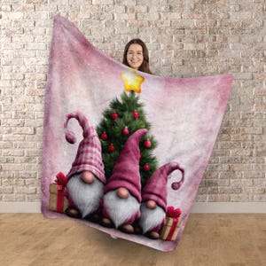 May include: A pink Christmas blanket with a watercolour design. It features three gnomes with pink hats and white beards, a decorated Christmas tree, and wrapped gifts. The blanket is held up against a brick wall.