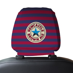 Op de afbeelding: Rode en blauwe gestreepte autostoel hoofdsteunhoes met een wit cirkelvormig logo met de tekst "Newcastle Brown Ale" met een blauwe ster en een skyline van de stad in de cirkel.