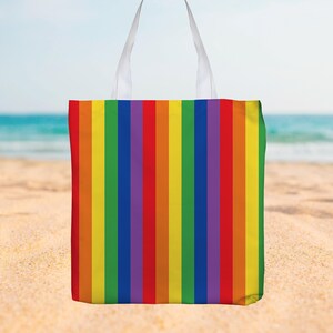 Op de afbeelding: Een draagtas met verticale strepen in regenboogkleuren: rood, oranje, geel, groen, blauw en paars. De tas heeft witte handvatten en staat op een zandstrand met de zee op de achtergrond.