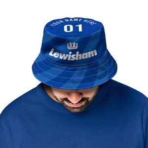 Może przedstawiać: Niebieski kapelusz typu bucket z białą koroną i nadrukiem "YOUR NAME HERE 01 Lewisham" z przodu.