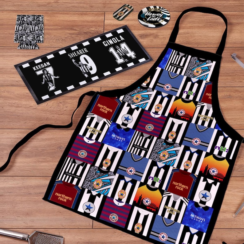 May include: A black apron with a patchwork design of soccer jerseys in various colors, including black and white stripes, blue, and red. A black and white rectangular mat with player names and numbers is also visible, along with coasters and a can cooler.