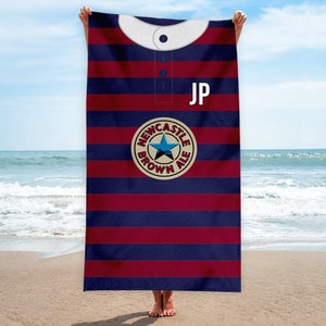 Peut inclure: Une serviette de plage ray&eacute;e bleu et bordeaux avec un col blanc et un logo circulaire repr&eacute;sentant une &eacute;toile et le texte "Newcastle Brown Ale". La serviette porte les initiales "JP" dans le coin sup&eacute;rieur droit.