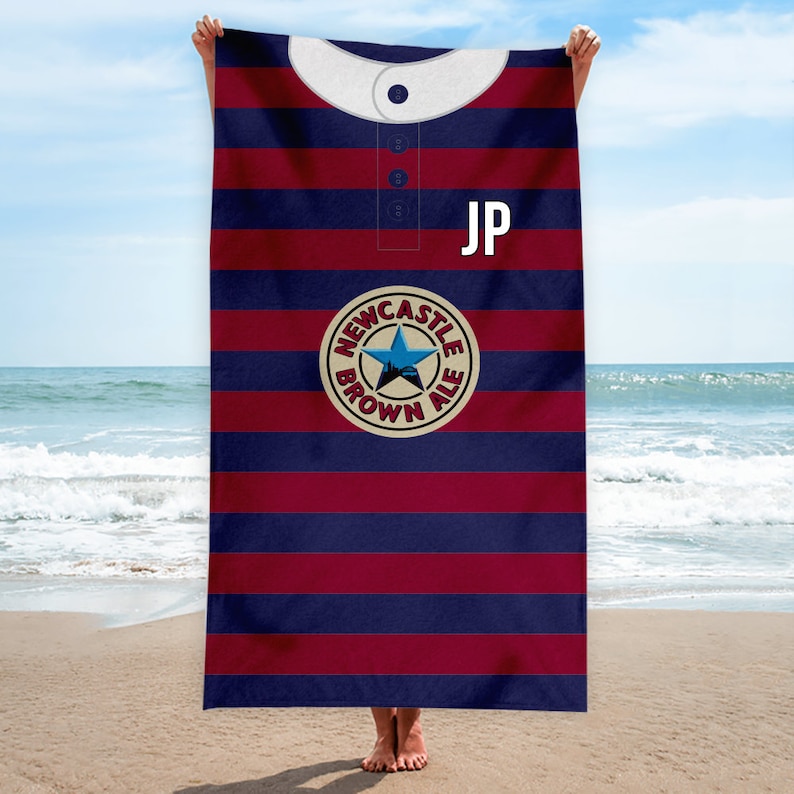 Peut inclure: Une serviette de plage ray&eacute;e bleu et bordeaux avec un col blanc et un logo circulaire repr&eacute;sentant une &eacute;toile et le texte "Newcastle Brown Ale". La serviette porte les initiales "JP" dans le coin sup&eacute;rieur droit.