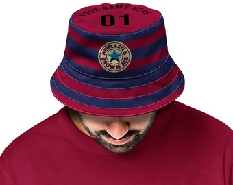 Puede incluir: Un sombrero de pescador a rayas rojas y azules con un logotipo de Newcastle Brown Ale y el número "01" en la parte delantera. El texto "Your Name Here" está impreso encima del número.