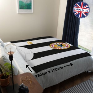 May include: A black and white striped blanket with a Newcastle United logo, measuring 59 inches by 59 inches. The blanket is displayed on a bed, showcasing the team's colors and emblem. The blanket has the text "JP" in gold.