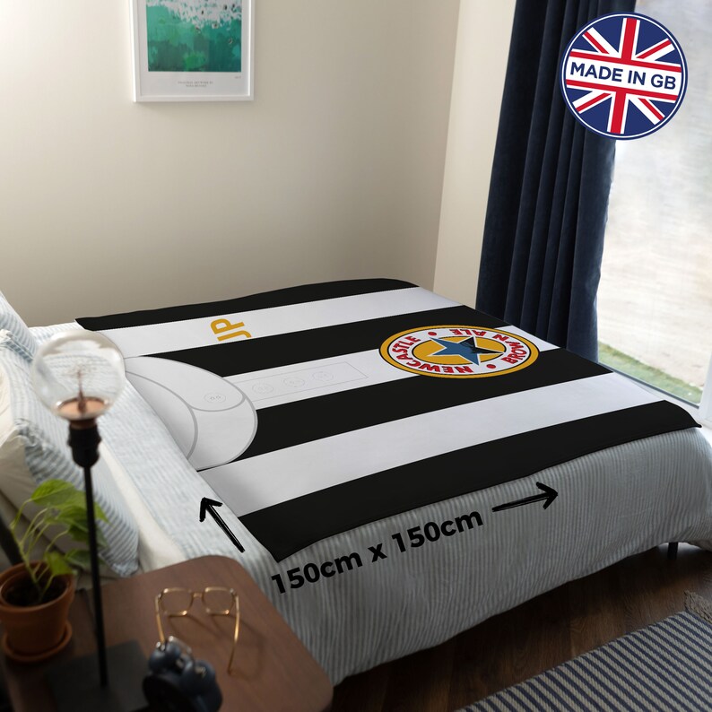 May include: A black and white striped blanket with a Newcastle United logo, measuring 59 inches by 59 inches. The blanket is displayed on a bed, showcasing the team's colors and emblem. The blanket has the text "JP" in gold.