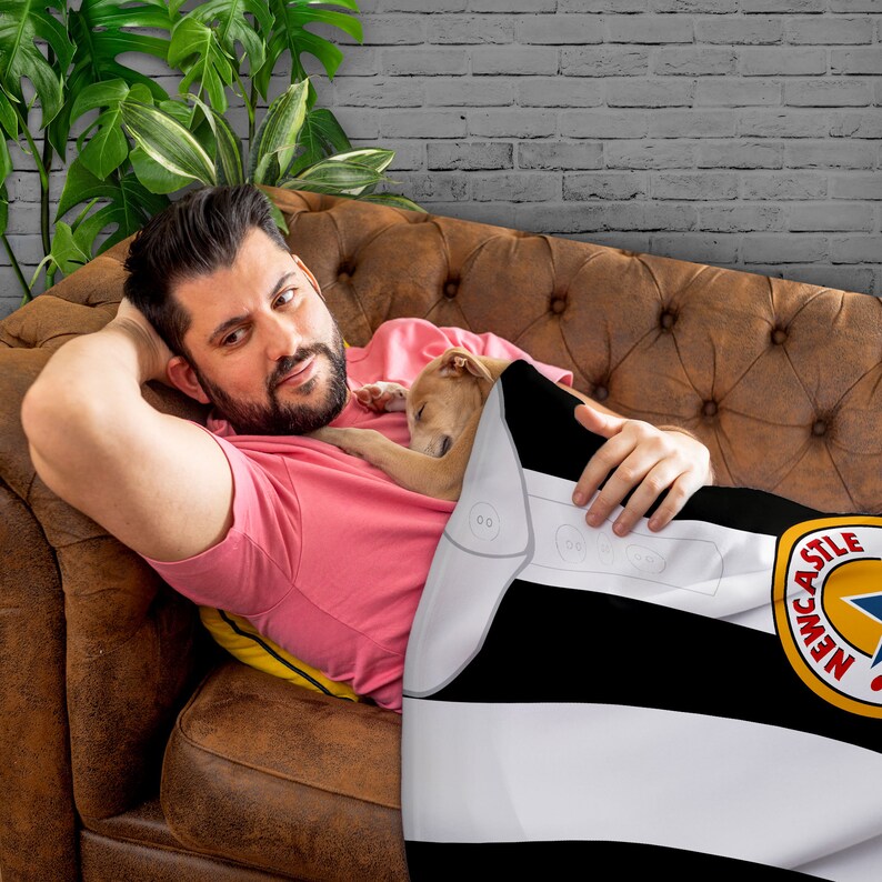 May include: A black and white striped blanket with the Newcastle United logo. The blanket has a white border with three small, round, gray buttons. The blanket is draped over a brown leather couch.