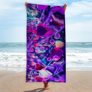 Könnte beinhalten: Ein lebendiges Strandtuch mit einem abstrakten Design in Lila-, Rosa- und Blautönen. Das Tuch weist geometrische Formen, Blasen und ein strukturiertes Muster auf. Das Tuch wird vor einem Strandhintergrund gehalten.