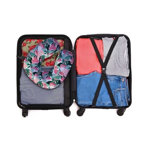 May include: A black suitcase with a pink and green floral print travel pillow inside. The suitcase is packed with clothes, including a red shirt, a striped shirt, and blue jeans.