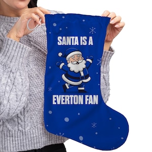 Puede incluir: Un calcetín navideño azul con el texto "SANTA IS A EVERTON FAN" y un dibujo animado de Papá Noel vestido de azul. El calcetín tiene copos de nieve blancos y un lazo plateado para colgar.
