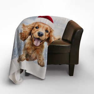 Può includere: Una coperta in pile bianco con un'illustrazione ad acquerello di un cocker spaniel dorato che indossa un cappello di Babbo Natale rosso. Il cane guarda la telecamera con la lingua di fuori. Lo sfondo è blu chiaro con fiocchi di neve bianchi.
