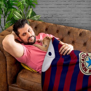 May include: A man and a small dog are lying on a brown couch. The man is wearing a pink shirt and the dog is white. They are both covered by a red and blue striped blanket with a white can design featuring the text "Newcastle" and a star.