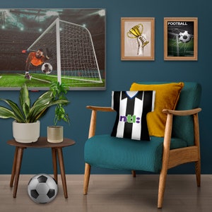 May include: A living room setting featuring a teal armchair, a side table, and a TV showing a football match. A black and white football shirt-shaped cushion is on the chair. A football rests on the floor. Two framed pictures are on the wall.