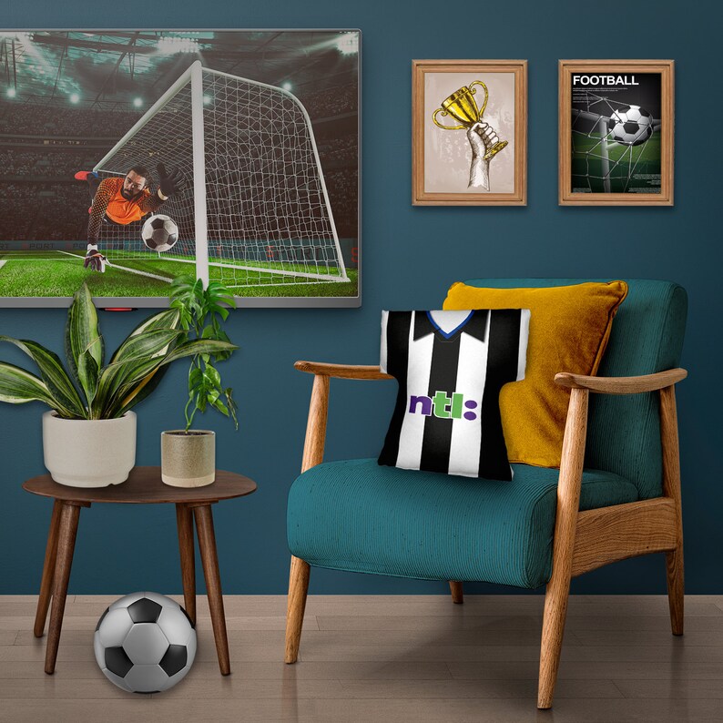 May include: A living room setting featuring a teal armchair, a side table, and a TV showing a football match. A black and white football shirt-shaped cushion is on the chair. A football rests on the floor. Two framed pictures are on the wall.