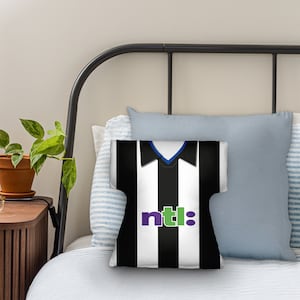 May include: A cushion shaped like a black and white striped football shirt. The shirt design features a blue collar and the word "ntl:" in purple and green. The cushion is placed on a bed with other pillows.