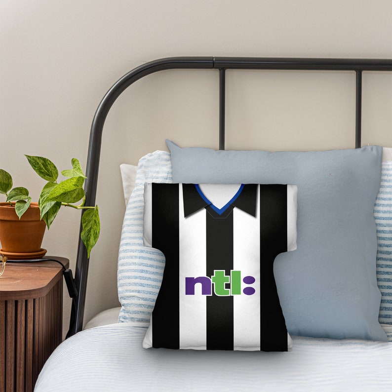 May include: A cushion shaped like a black and white striped football shirt. The shirt design features a blue collar and the word "ntl:" in purple and green. The cushion is placed on a bed with other pillows.