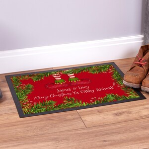 May include: A red Christmas doormat with a green pine garland border. The doormat features two elf legs wearing red and white striped socks and green elf shoes. The text on the doormat reads "James & Lucy Merry Christmas Ya Filthy Animal!"