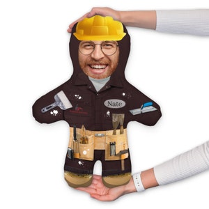 May include: A brown plush toy shaped like a person wearing a yellow hard hat, brown work overalls, and a tool belt. The toy has a smiling face with glasses and the name "Nate" on the chest.