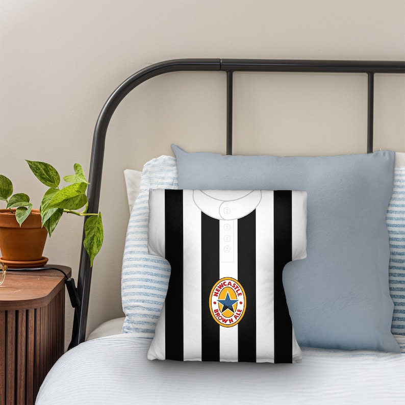 May include: A black and white striped throw pillow with a Newcastle Brown Ale logo. The pillow is on a bed with a blue and white striped pillow behind it.