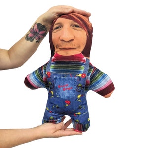 Può includere: Un giocattolo di peluche con la faccia di una persona stampata su di esso. Il giocattolo indossa una camicia a righe blu e rosse, una salopette blu con una toppa "Good Guys" e un berretto da baseball rosso.