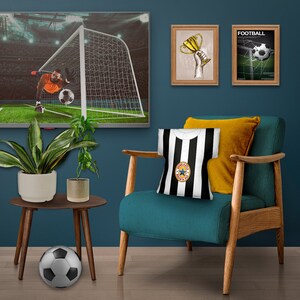 May include: A teal armchair with wooden arms and legs, a black and white striped throw pillow with a star logo, and a yellow throw pillow. The chair is in front of a blue wall with two framed pictures of a trophy and a football game.